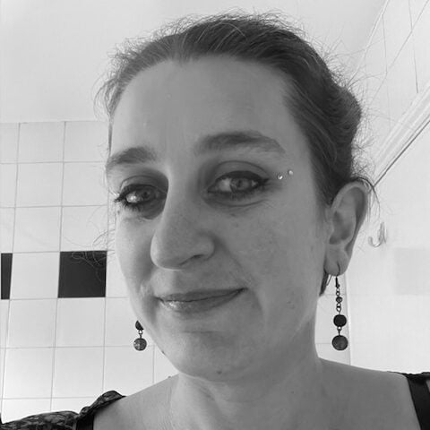 A black and white headshot of Emily Jenkins, a white woman with brown hair and beaded earrings, smiling at the camera in front of a tiled wall in the background.