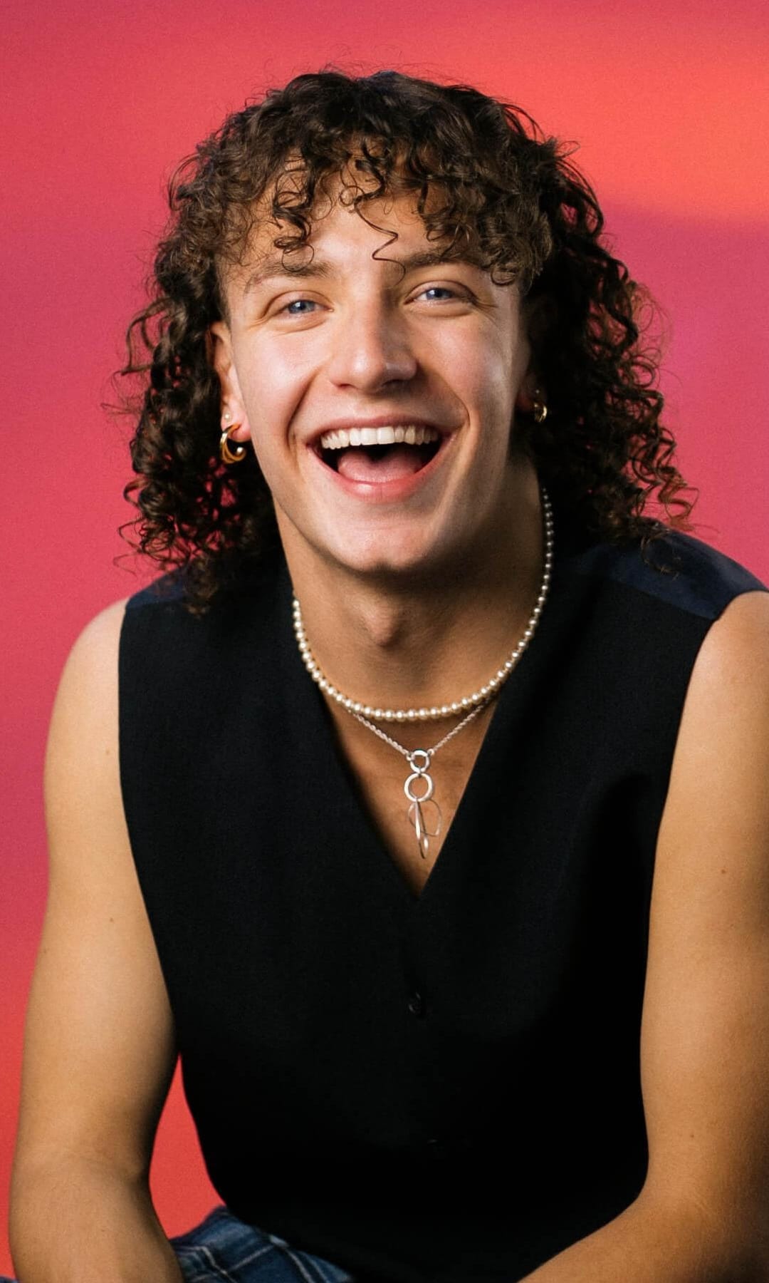 Rhys Wilkinson A headshot of actor Rhys Wilkinson. They are sitting and facing the viewer, laughing with shoulder length brown curly hair. They're wearing a pearl necklace and a black sleeveless tank top.